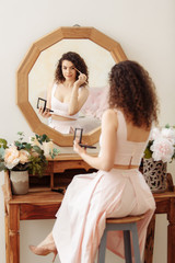 Young happy girl with curly hair does makeup in front of a vintage mirror. A beautiful woman in a pink dress paints her eyes.