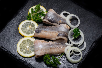 Salted Deboned Herring Fillets garnished with Yellow Onion, Lemon and Fresh Parsley. Natural black stone background. Clupea harengus.