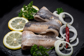 Salted Herring Fillets garnished with Yellow Onion, Lemon, Fresh Parsley and Dried Chili Pepper. Natural black stone background. Clupea harengus.