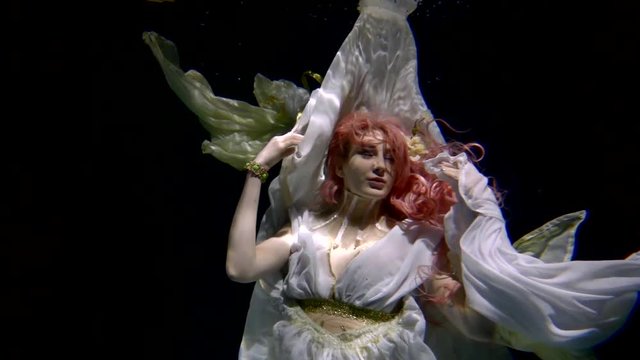 smiling magic princess is plunging inside water of sea in night, looking at camera, artistic underwater shot