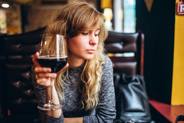 A woman in a gray top is sitting alone in a restaurant with a glass of wine. Romantic girl with blonde curls, relaxing in a cafe and shy smile, looking down.