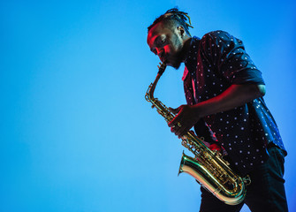 Young african-american jazz musician playing the saxophone on blue studio background in trendy neon...