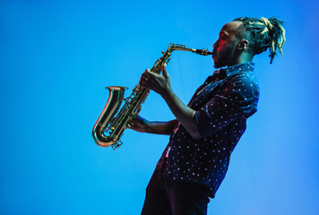 Young african-american jazz musician playing the saxophone on blue studio background in trendy neon...