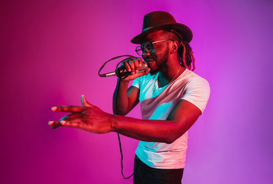 Young african-american jazz musician with microphone singing a song on purple studio background in trendy neon light. Concept of music, hobby, inspirness. Colorful portrait of joyful attractive artist
