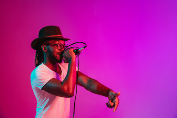 Young african-american jazz musician with microphone singing a song on purple studio background in trendy neon light. Concept of music, hobby, inspirness. Colorful portrait of joyful attractive artist