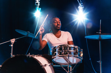 Young african-american jazz musician or drummer playing drums on blue studio background in glowing smoke around him. Concept of music, hobby, inspirness. Colorful portrait of joyful attractive artist.
