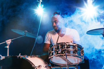 Young african-american jazz musician or drummer playing drums on blue studio background in glowing smoke around him. Concept of music, hobby, inspirness. Colorful portrait of joyful attractive artist.
