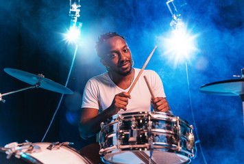 Young african-american jazz musician or drummer playing drums on blue studio background in glowing...