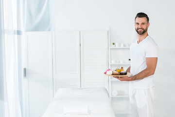cheerful bearded man holding tray with bottles of oil, bowls with sea salt and flowers in spa center