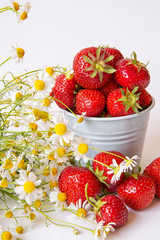 Scattered strawberry berry and in a small bucket on a light background with a bouquet of chamomile flowers . Summer berry season.