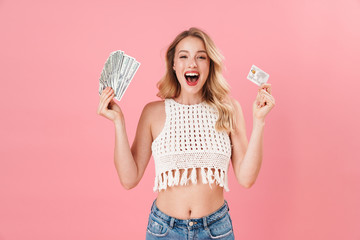Happy young woman posing isolated over pink wall background holding money and credit card.