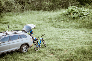 Professional cyclist resting with bicycle while sitting in the car trunk on the green meadow in the...