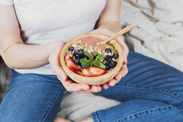 Closeup of woman in jeans and white shirt in her bed holding vegan smoothie bowl, selective focus