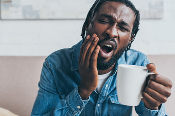 young african american man holding coffee cup while suffering from toothache