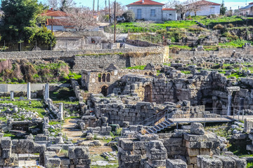 Excavated archaeological ruins in Ancient Corinth Greece with stairs and walkway for tourists and red tile roofed village houses behind