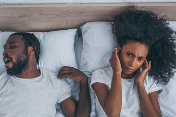top view of african american woman suffering from headache while lying in bed near snoring husband
