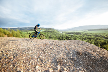 Fototapeta premium Professional well-equipped cyclist riding bicycle on the rocky mountains with beautiful landscape view during the sunset. wide view with copy space