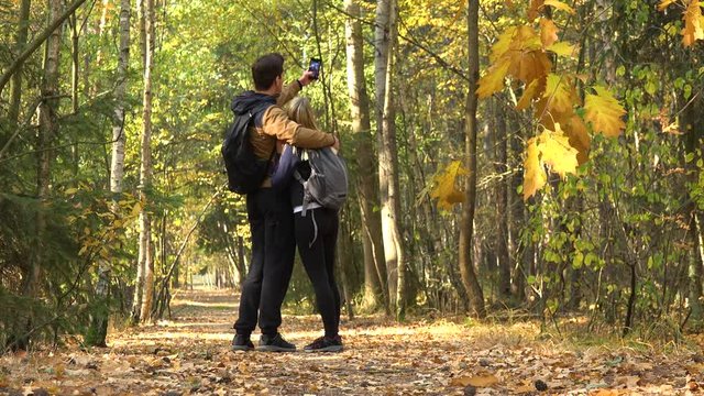 A hiking couple walks through a forest on a sunny day, stops to take a selfie with a smartphone and walks on