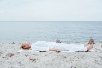 attractive blonde woman with closed eyes meditating while lying on yoga mat near sea