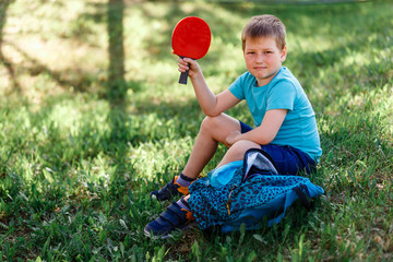 an eight-year-old boy sits on the green grass and holds a table tennis racket in his hands