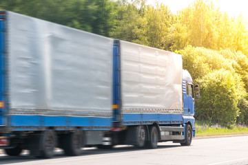 Blue delivery van and white truck driving on the asphalt road between line of trees in autumn landscape