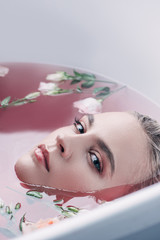Fototapeta premium attractive young woman lying in bathtub with colored pink water and flowers and looking at camera