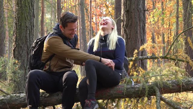 A hiking couple sits on a broken tree in a forest and tickles each other