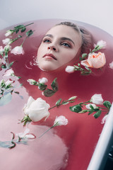 beautiful young woman lying in bathtub with colored pink water and flowers and looking away