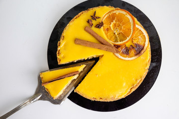 top view of a round cake of yellow-orange color decorated with a slice of orange and cinnamon sticks one piece of which lies on the blade cut