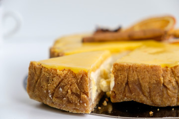 round cheesecake yellow-orange decorated with cinnamon sticks and orange slices from which beautifully cut off a triangular piece on a white background