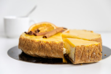 round cheesecake yellow-orange decorated with cinnamon sticks and orange slices from which beautifully cut off a triangular piece on a white background, where the background is visible blurred mug