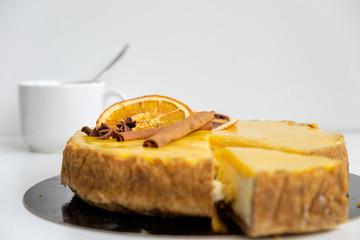 round cheesecake yellow-orange decorated with cinnamon sticks and orange slices from which beautifully cut off a triangular piece on a white background, focus on cake decoration