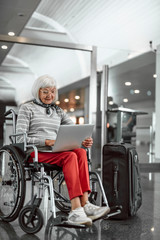Fototapeta na wymiar Happy old lady on disabled carriage typing at laptop at airport