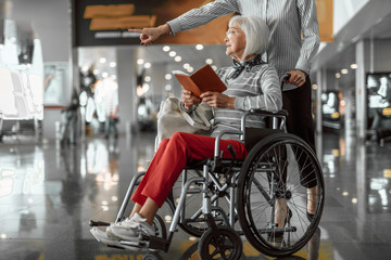 Female worker of airport staying with elderly woman in wheelchair at hall