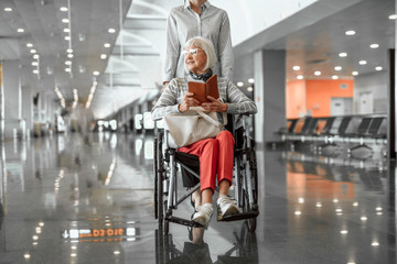 Young female worker of airport holding wheelchair with elderly lady in hall