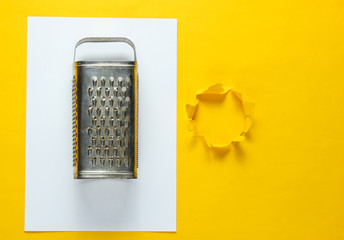 Old metal grater on yellow paper background with torn hole for copy space. Top view. Studio shot