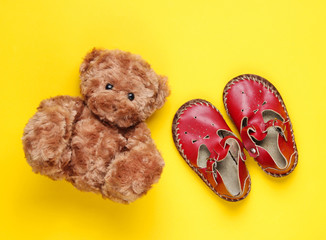The concept of childhood. Red leather baby sandals, teddy bear on a yellow background. Top view
