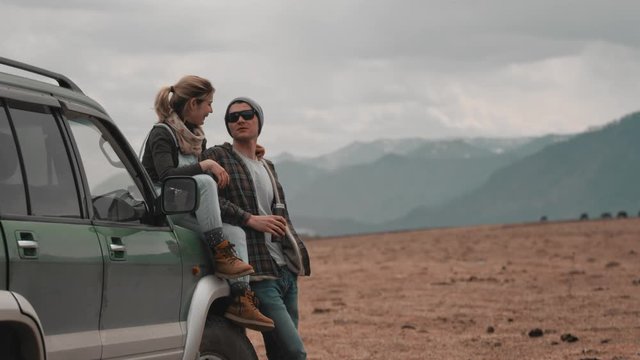 A loving couple of travelers at the SUV in the valley during a summer trip. A woman sitting on the hood of a car, a man drinking from a thermal mug, talking, kissing. On a halt. Slow motion, 4K