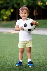Portrait of little toddler child standing in green football field holding soccer ball. Smiling little football player at stadium. Future footbal star and active childhood concept