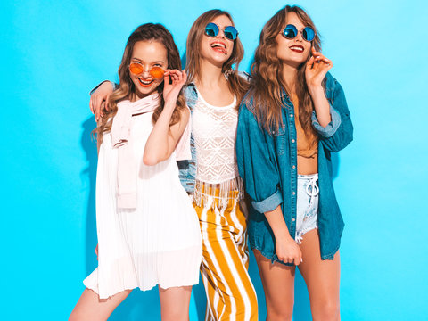 Three young beautiful smiling hipster girls in trendy summer casual clothes. Sexy carefree women posing near blue wall. Positive models going crazy in sunglasses