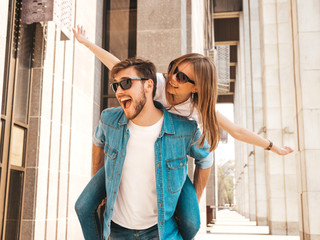 Smiling beautiful girl and her handsome boyfriend in casual summer clothes. Man carrying his girlfriend on the back and she raising her hands.Happy cheerful family having fun on the street background