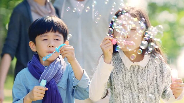 asian little girl and boy brother and sister blowing bubbles in park with parents watching from behind