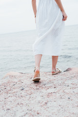 cropped view of woman in white dress and sandals standing near sea