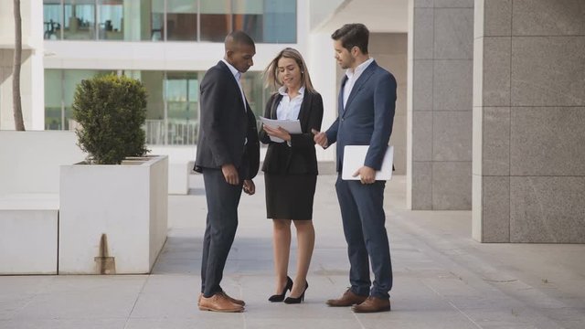 Multiethnic business people discussing papers outdoor. Professional young businessmen and businesswoman with documents and laptop standing outside modern office building. Business concept