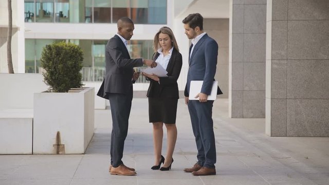 Young multiethnic business people discussing papers outdoor. Professional businessmen and businesswoman with documents and laptop standing together outside modern office building. Business concept