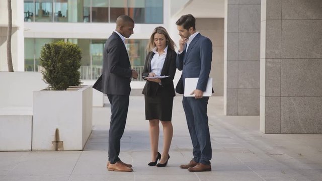 Male and female coworkers discussing papers outdoor. Young multiethnic business colleagues with laptop and documents standing together outside modern office building. Business concept