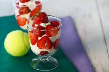 Whipped cream and strawberries served in a glass. Purple and dark green napkins, white wooden table, tennis ball, high resolution