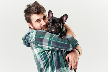 Hipster Bearded guy holding and hugging a nice French Bulldog dog in his arms with love and playing with him, against a white background
