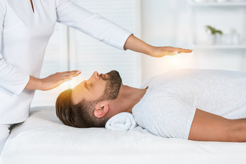 cropped view of healer putting hands above head while healing handsome man on massage table