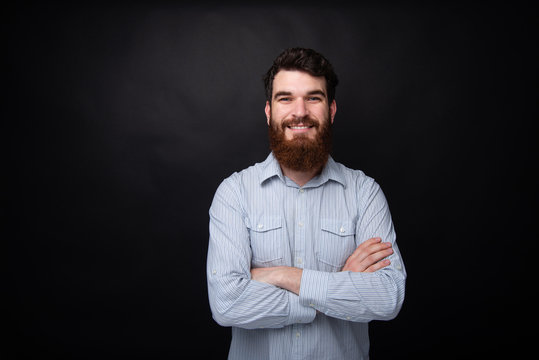 Portrait of Cheerful bearded man smiling with crossed arms over dark isolated gray background
